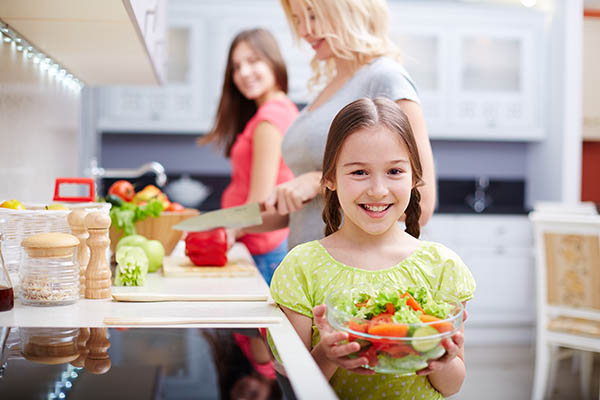 Portrait of happy girl with vegetable salad on background of her mother and sister cooking